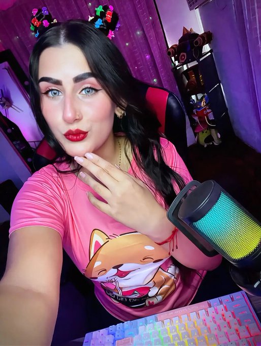A woman with dramatic makeup and red lipstick sits in a gaming chair, wearing a pink t-shirt featuring an anime character, with a colorful RGB microphone and keyboard visible.