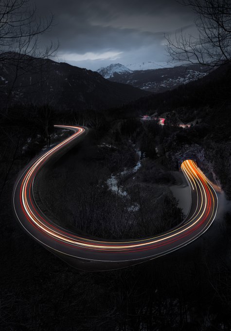 A long-exposure photograph of a winding mountain road at night, featuring light trails from vehicles curving through a dark, forested landscape with snow-capped mountains visible in the distance.