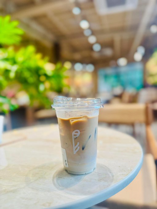 A clear plastic cup containing an iced coffee drink with ice and what appears to be condensed milk, placed on a marble table inside a café with blurred greenery and seating in the background.