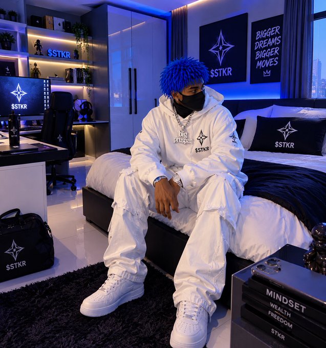 A person sitting on a bed in a blue-lit room, wearing a white outfit, a blue fluffy wig, and a black mask, with 'S$TKR' branding visible on pillows, decor, and books.