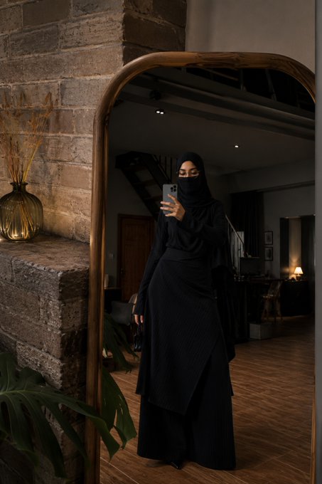 A woman wearing a black niqab and long black pleated dress takes a mirror selfie with her phone in a room featuring exposed brick walls, a large arched mirror, and warm ambient lighting.