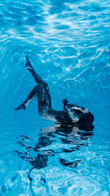 An underwater photograph of a person in a black bikini diving headfirst into a bright blue pool, with reflections visible on the water's surface.