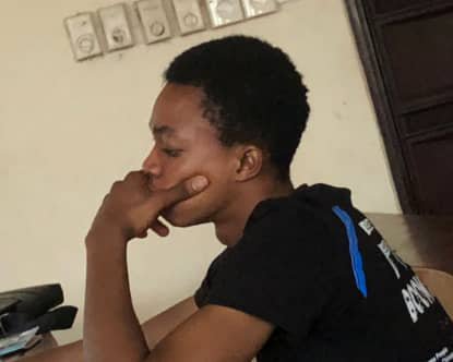 A young man in a black shirt rests his chin on his hand in a thoughtful pose inside a room.