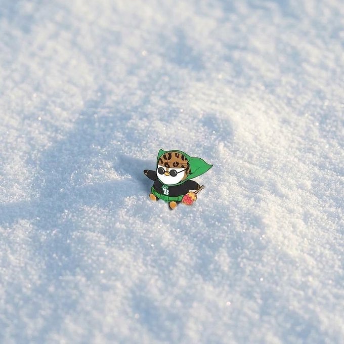 An enamel pin depicting a small character with a leopard-print hat and green outfit sits on a snowy surface.