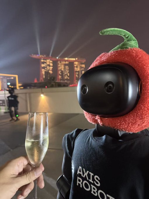 A robot wearing an 'AXIS ROBOT' shirt and a red strawberry-like hat with a green leaf holds a champagne glass, with the illuminated Marina Bay Sands hotel visible in the night background.