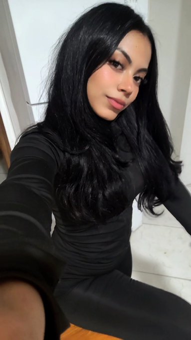 A selfie of a woman with long black hair wearing a black turtleneck and dark pants, posing indoors.