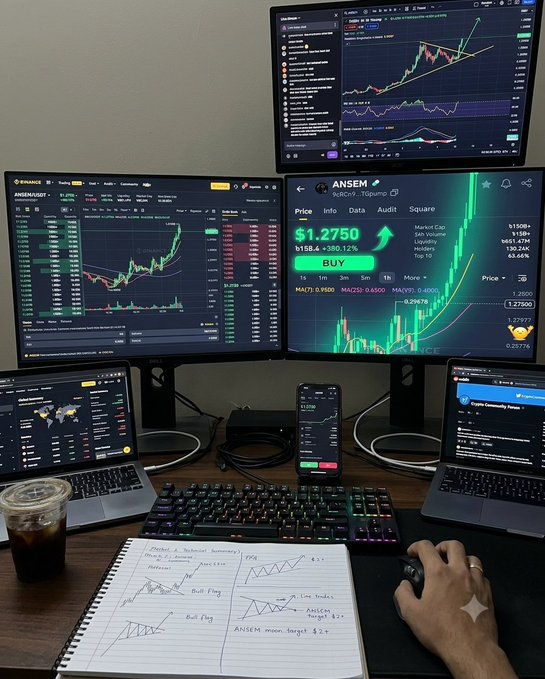 A cryptocurrency trading setup with multiple monitors displaying charts for ANSEM/USD, a notebook with technical analysis notes including 'Bull Flag' and 'ANSEM', and a hand on a mouse next to an iced coffee.