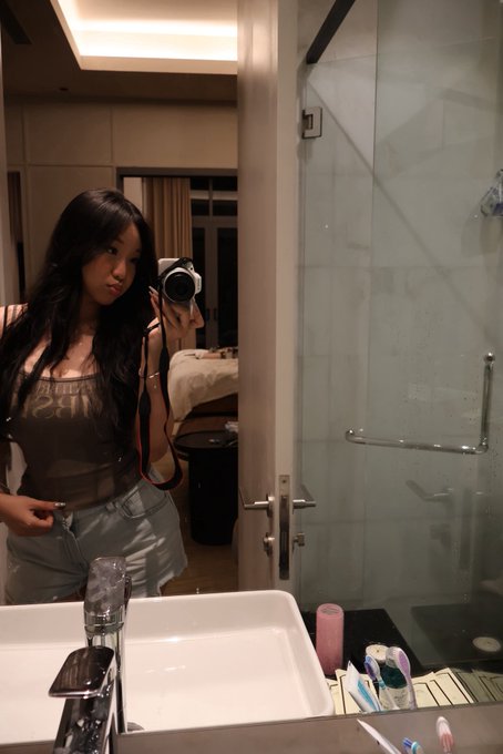 A mirror selfie of a woman holding a camera in a bathroom, wearing a brown top and light blue jeans.
