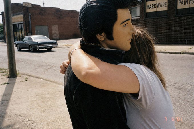 A vintage film still showing Elvis Presley in a black leather jacket embracing a woman on a street, with a classic car and 'HARDWARE PAINTS' storefront in the background.