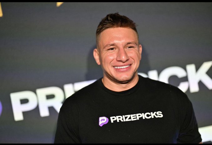 A smiling man wearing a black shirt with the 'PRIZEPICKS' logo stands in front of a backdrop displaying the same brand name.