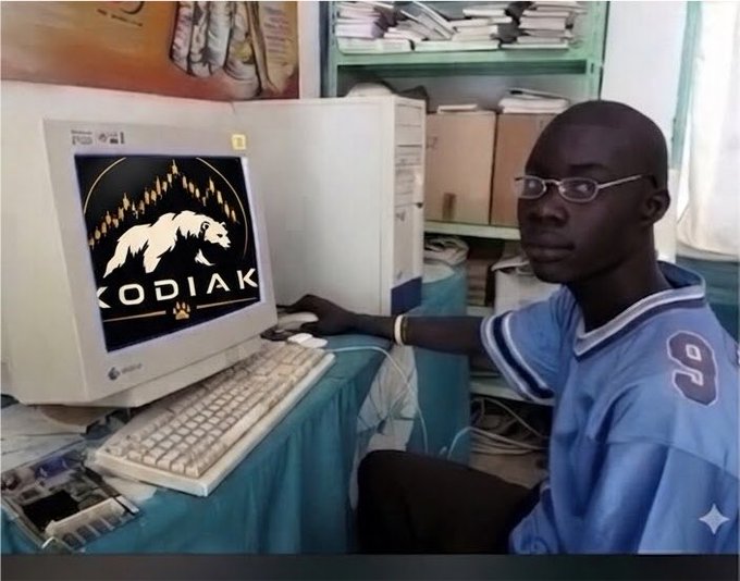 A young man sits at an older desktop computer displaying a "KODIAK" logo featuring a bear graphic on the monitor, with shelves of files visible in the background. The image visually represents the Kodiak platform referenced in the post, suggesting a user engaged with the service to find web3 work opportunities.