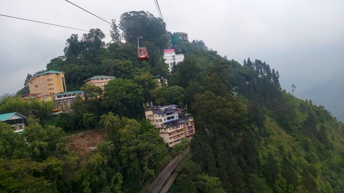 An aerial view shows a lush green hillside with buildings, including hotels and a cable car with a red gondola traveling along wires above the trees.