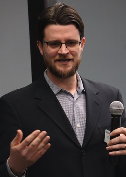 The image shows a young man with glasses, a beard, and brown hair, wearing a dark blazer over a light checkered shirt, holding a microphone while gesturing with his other hand during what appears to be a speaking engagement.

The accompanying text relates to the image by identifying the man as William Lawrence, a Democratic Socialist projected to win a Congressional seat, whose past radical positions on abolishing police, prisons, and the Senate are being highlighted in the post.