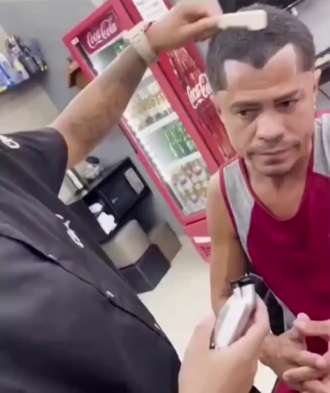 The image shows a man getting his hair cut by another person, who appears to be a barber or hairstylist. The man receiving the haircut has a shaved head with a noticeable mohawk style. Both individuals are wearing black shirts, which contrasts with their surroundings. In the background, there is a Coca Cola vending machine and a refrigerator, indicating that they might be in a store or salon setting. A person's hand can also be seen holding a cell phone, possibly capturing the moment or looking up information related to the haircut process. The image includes text that reads "WATCH: Monero gang member gets his initiation but doesn't like the result much." This suggests that there is an ongoing conversation between the two individuals in the scene.