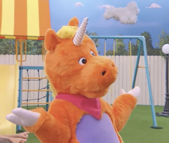 A costumed orange unicorn character from a children's show standing in a colorful playground set with a swing set and fence in the background.