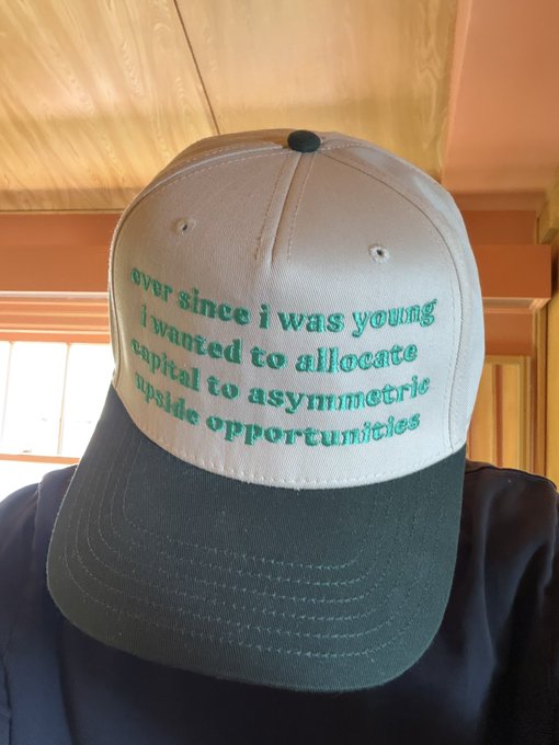 A white and dark green snapback cap embroidered with the green text "ever since i was young i wanted to allocate capital to asymmetric upside opportunities," worn by a person, playfully personifies the @base post's message about seeking asymmetric upside opportunities by suggesting the cap itself has always aspired to allocate capital to such ventures.