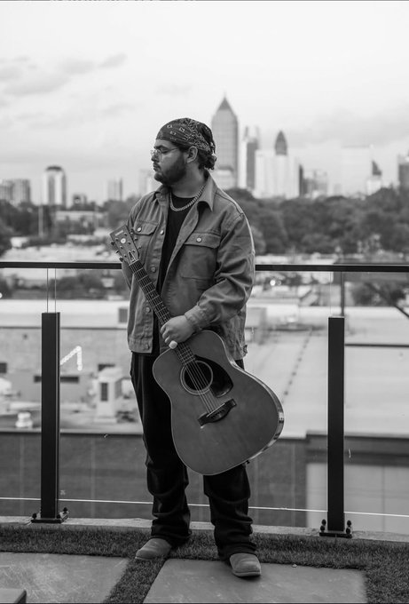 A black-and-white photograph of a bearded man wearing a bandana and jacket, holding an acoustic guitar on a balcony overlooking a city skyline.