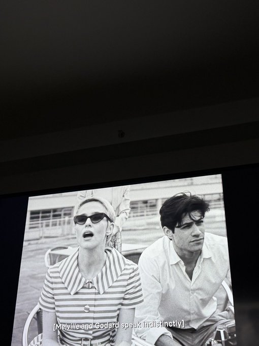 A photo taken of a cinema screen displaying a black and white film scene with two people, captioned '[Melville and Godard speak indistinctly]'.