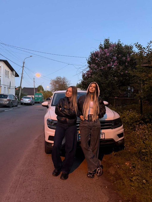 Two women posing in front of a white SUV on a street at dusk, both wearing dark outfits with one in a black leather jacket and the other in a leather jacket with a hood.