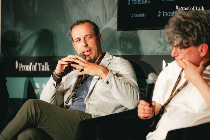 The image shows a man sitting down with his mouth open while another person sits next to him. They are both holding microphones, suggesting that they might be participating in an event or discussion where they are speaking into them. The background features a green wall with a large screen displaying information about TACOS and other topics related to the event.
