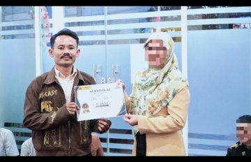 A photograph of an award ceremony where a man in a brown jacket presents a certificate to a woman in a hijab; some faces in the background are blurred for privacy.