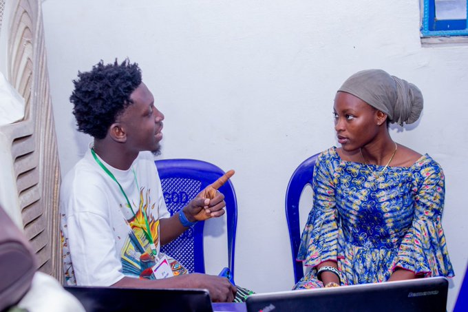 Two people seated in blue plastic chairs having a conversation; the man on the left wears a lanyard and points toward the woman wearing a headscarf and blue patterned dress.