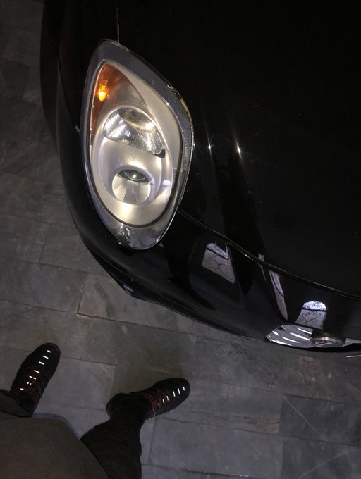 A nighttime photo taken from above shows a person's sneakers next to a black car with its headlight turned on, reflecting light off the polished hood. The illuminated headlight complements the casual, carefree vibe of the post wishing followers a blessed night.