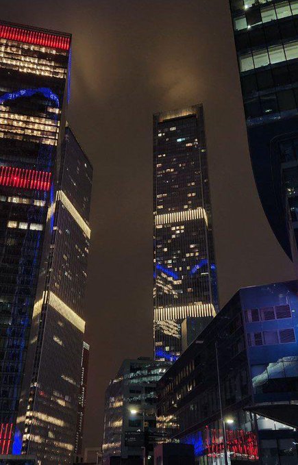 A nighttime cityscape photo featuring a tall illuminated skyscraper and adjacent modern buildings glowing with blue and red lights against a dark sky.