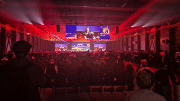 The image shows a large crowd of people gathered for an event, with some sitting in chairs while others stand around watching a presentation or performance. The stage has a screen displaying information about Bitcoin, possibly related to an art exhibition showcasing various cryptoart pieces. The audience appears engaged and interested in the content being presented.