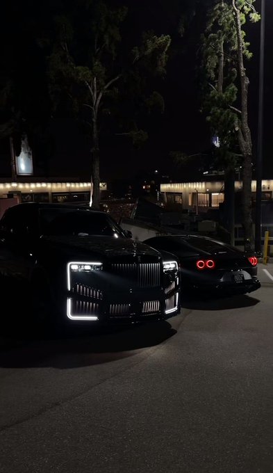 The image shows two luxury cars parked side by side, with a building visible behind them. The cars are positioned next to each other, creating an interesting contrast between their sleek designs and the surrounding environment. The scene appears to be set at night, as indicated by the dark background and the presence of streetlights in the vicinity.