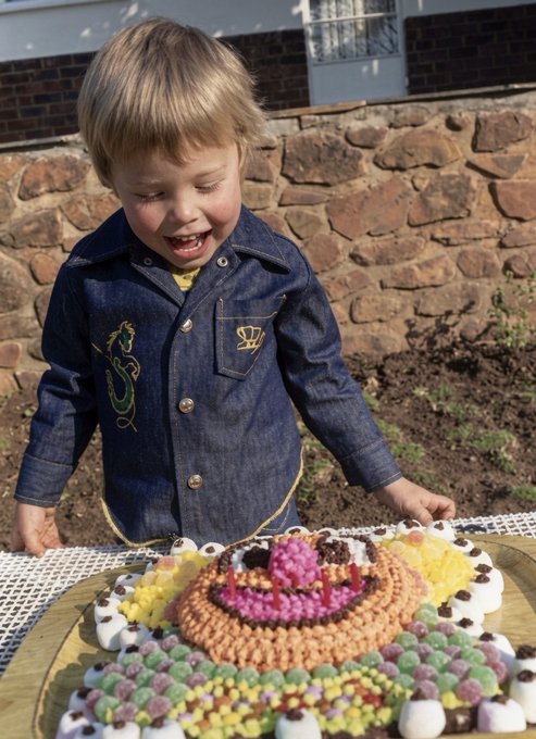 A young boy in a denim shirt reacts with excitement as a birthday cake decorated with colorful candies and lit candles sits before him on an outdoor table.