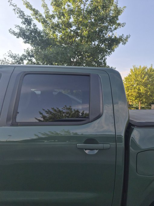 A green pickup truck's rear cab window reflects a clear blue sky, with the door handle below—nothing else visible. No crypto-related text, tickers, or signs appear in the image.