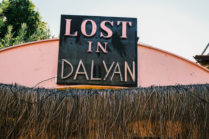 A black sign with white and pink lettering reading 'LOST IN DALYAN' mounted on a pink wall above a thatched roof.
