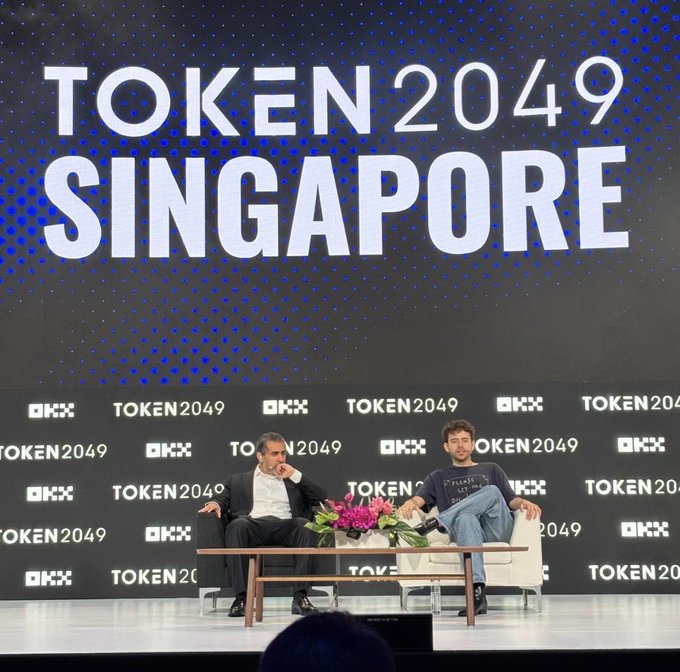 Two men engaged in conversation on a stage at the TOKEN 2049 SINGAPORE conference, seated in white chairs against a backdrop branded with 'OKX' and 'TOKEN 2049'.