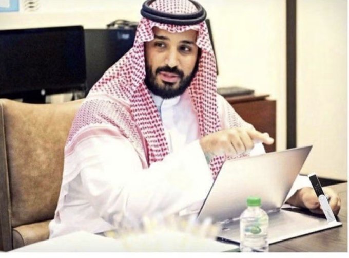 Mohammed bin Salman, Crown Prince of Saudi Arabia, wearing traditional dress and keffiyeh, seated with a laptop.