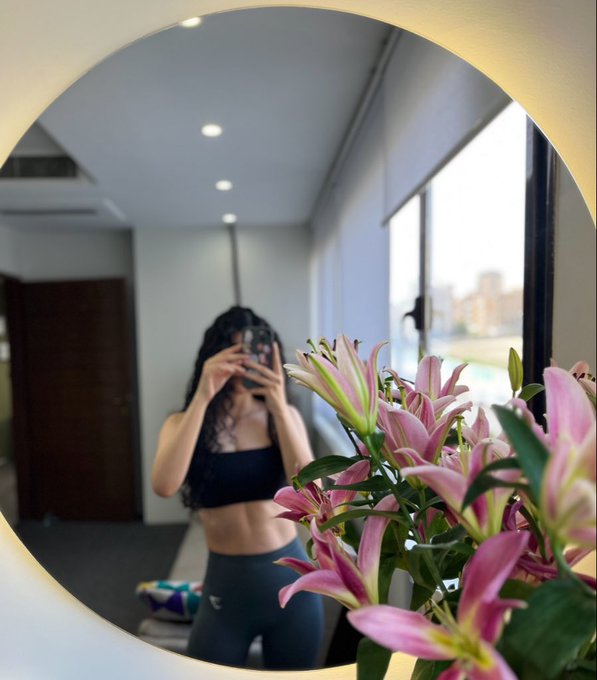 A mirror selfie of a woman in workout attire holding a camera, with a vase of pink lilies in the foreground and a window view in the background.