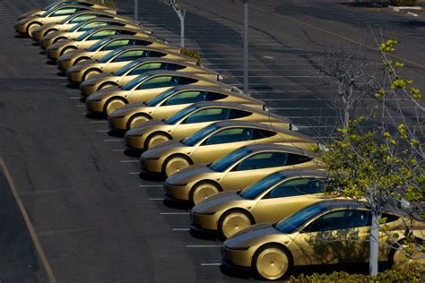 An aerial view of a parking lot filled with a diagonal row of identical gold-colored cars, with some green foliage visible on the right side.