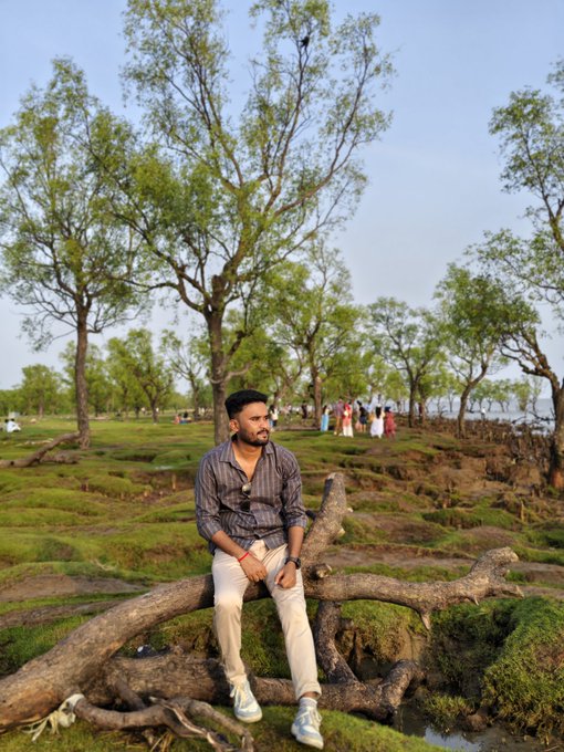 The image features a man sitting on a fallen tree branch, surrounded by trees and grass. The man appears to be wearing a striped shirt and white pants while he sits there. In the background, there are other people present as well. The photo is related to an advertisement for a sleep app called @useTria Community-powered growth with @River4fun Big airdrop coming soon, which encourages users to spend more time on their phones instead of engaging in other activities or getting proper rest.