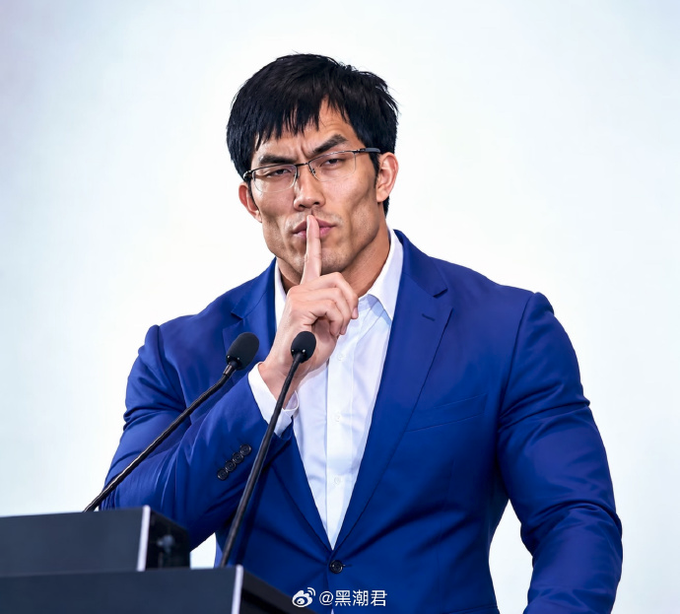 A man in a blue suit and glasses pressing his finger to his lips in a 'shush' gesture while standing at a podium with two microphones, with the watermark '@黑潮君' visible at the bottom.