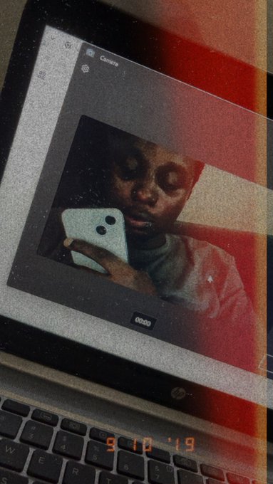 An artistic double-exposure style image showing a laptop screen displaying a person holding a phone in the camera app, with red lighting and the date "9.10.19" on the keyboard.