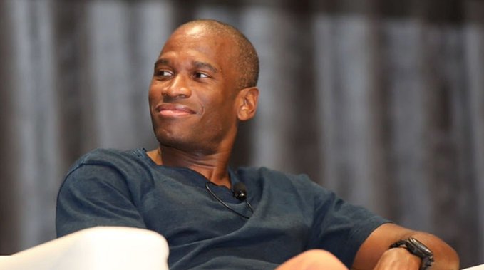 A photograph of a smiling bald man in a dark t-shirt sitting against a gray curtain backdrop.