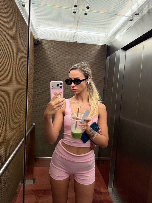 A mirror selfie of a young woman with blonde hair wearing dark sunglasses, a pink crop top, and pink shorts, holding a pink iPhone and an iced beverage with a straw, with AirPods in.