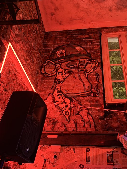 A red-lit brick wall features a mural of a cartoon monkey wearing a hat and smoking a cigar, with a neon triangle light and a window visible.