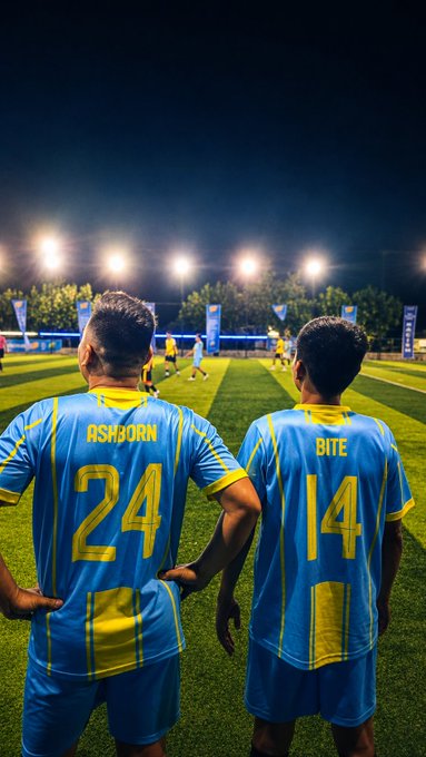 Two soccer players stand with hands on hips on a green field, wearing blue and yellow jerseys with the names "ASHOORN" (number 24) and "BITE" (number 14), illuminated by stadium lights at night.