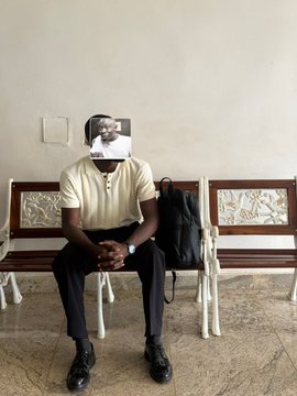 A man in a yellow polo shirt and dark trousers sits on a bench, holding a small photograph of himself over his face.