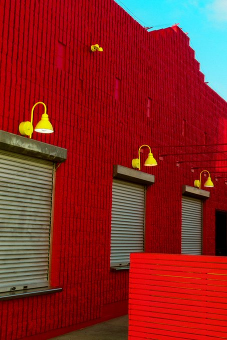 A striking red textured wall adorned with three vibrant yellow lamps above closed metal roll-up shutters, accompanied by a red bench in the foreground. The image showcases the artwork titled "Red Wall" by @CinziaGabrielPH, part of the #WALLS drop.