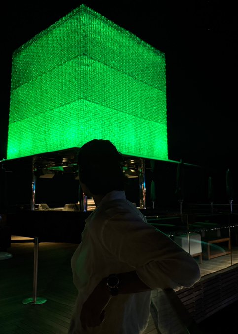 A person in a white shirt stands silhouetted in the foreground, gazing up at a large, cube-shaped structure illuminated in bright green at night.