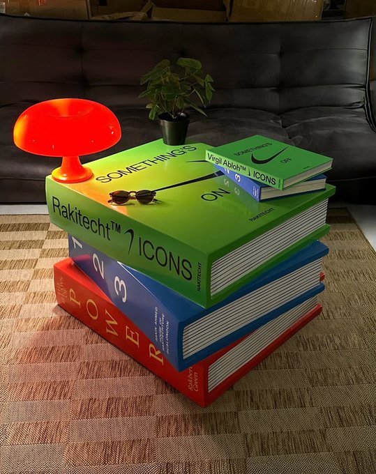 A stack of three oversized decorative books labeled 'SOMETHINGS', 'Rakitect™ 7 ICONS', and 'TRENDS' sits on a textured rug, topped with a green plant and an orange lamp.