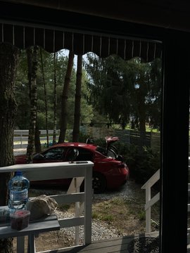 A view from a shaded porch looks out onto a red sedan parked in a gravel driveway surrounded by tall trees, with a water bottle resting on the railing. The image humorously supports the "touching grass" post by showing a glimpse of the outdoors waiting beyond the comfort of the covered deck.