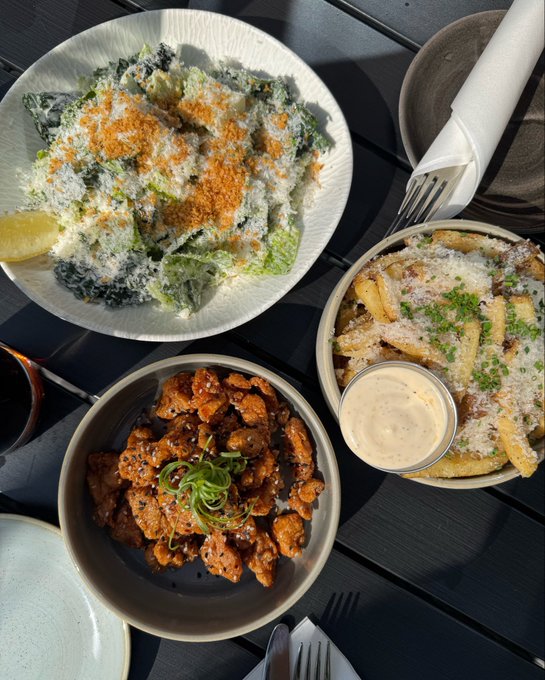 A Caesar salad, garlic parmesan fries with dipping sauce, and a bowl of glazed boneless wings arranged on a dark table with cutlery. The "girl dinner" spread plays on the post's humor, presenting a typical casual meal as if evaluating a crypto trade under $1 SOL.